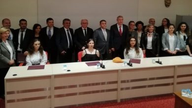 Görselde, bir masanın arkasında oturan üç kadın yer alıyor. Kadınların arkasında ise, ayakta duran kadın ve erkeklerden oluşan kalabalık bir grup bulunuyor.