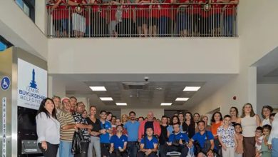 Görselde, İzmir Büyükşehir Belediyesi'ne ait bir binada çekilmiş toplu bir fotoğraf yer alıyor. Alt katta çeşitli yaş ve engel grubundan bireyler ile onların aileleri ve destekçileri bir araya gelmiş. Birkaç kişi tekerlekli sandalye kullanıyor. Arka planda “İzmir Büyükşehir Belediyesi” yazılı bir engelli aracı şarj istasyonu dikkat çekiyor. Üst katta ise kırmızı tişört giymiş kalabalık bir grup, balkonun arkasından aşağıdaki grubu izliyor.