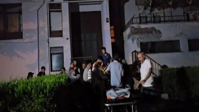 Görselde, gece vakti bir apartman önünde toplanmış bir grup insan görülüyor. Ortam karanlık ve acil bir durum izlenimi veriyor. Ön planda boş bir sedye yer alırken, arka planda sağlık ve kurtarma ekipleri ile bazı vatandaşlar bir kişiye müdahale ediyor gibi görünüyor. Bina eski ve hasarlı bir yapı izlenimi veriyor.