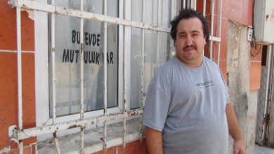 Görselde, bir apartman dairesinin önünde gri tişörtlü bir adam (engelli birey Gökhan Bürelli) kameraya doğru bakarak ayakta duruyor. Arkasında, demir parmaklıklı pencerenin camında siyah harflerle “Bu evde mutluluk var” yazısı dikkat çekiyor. Ancak pencere parmaklıkları eğilmiş, camlarda çatlak izleri var; bu da yaşanan rahatsızlıkların izlerini taşıyor.