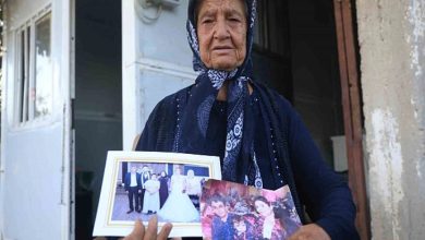 Bu görselde, başörtülü yaşlı bir kadın, elinde iki aile fotoğrafı tutarak kameraya bakıyor. Kadının yüz ifadesi hüzünlü ve derin bir acıyı yansıtıyor. Arka planda mütevazı bir evin girişi görülüyor. Fotoğraflardan biri düğün anına, diğeri ise ailece çekilmiş bir kareye ait.