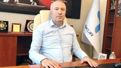 Görselde, bir makam odasında oturan bir erkek yer alıyor. Kendisinin ciddi ve kararlı bir ifadesi var. Üzerinde açık mavi bir gömlek bulunuyor. Masasının arkasında Türkiye İş Kurumu'na (İŞKUR) ait bir bayrak, çeşitli plaketler ve ödüller yer almakta. Sol arka planda Atatürk'ün yer aldığı bir tablo dikkat çekiyor. Masa üzerinde bir su şişesi ve çerçeveli bir fotoğraf da görülüyor.