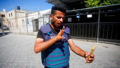 Görselde, Gazze’de bulunan bir erkek aktivist, üzerinde mavi koruyucu basın yeleğiyle cep telefonuna bakıyor ve işaret diliyle iletişim kuruyor. Arka planda basit yapılar, tellerle çevrili bir alan ve açık hava ortamı görülüyor. Güneşli bir günde çekilmiş olan bu karede, kişi kararlı bir ifadeyle dünyaya mesajını iletmeye çalışıyor.