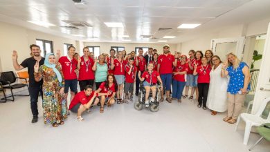 Bu görselde, geniş ve aydınlık bir kapalı mekânda toplanmış kalabalık bir grup insan görülüyor. Çoğu kişi kırmızı tişört giymiş ve boynunda beyaz fular takmış, bu da muhtemelen bir organizasyona veya etkinliğe ait olduklarını gösteriyor. Ortada tekerlekli sandalyede bir birey bulunuyor. Sol köşede geleneksel kıyafetli bir kadın ve sağ köşede mavi puantiyeli bluz giymiş bir kadın yer alıyor.