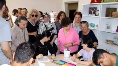 Görselde, bir masanın etrafında oturan engelli bireyler kağıt üzerine resim yapıyor. Engelli bireylerin çevresini ise yaşça büyük çoğunluğu kadın olan bir grup olduğu görülüyor.