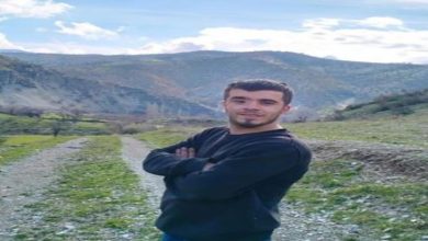 Görselde, doğa manzaralı bir kırsal alanda genç bir erkek kamera karşısında poz veriyor. Kollarını göğsünde kavuşturmuş, gülümsüyor. Üzerinde siyah bir kazak ve mavi pantolon var. Arka planda dağlar, tepeler ve açık mavi gökyüzü dikkat çekiyor. Zemin taşlık ve otlarla kaplı bir patika yolu gösteriyor.