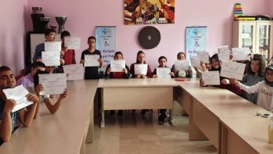 Görselde, bir sınıf ortamında "sertifika töreni" havası yansıtılıyor. U şeklinde yerleştirilmiş masaların etrafında oturan 15'e yakın genç birey, ellerinde sertifikalarını gururla tutuyor. Arkada iki afiş yer alıyor: Üzerinde “İş Kulübü” ve “Bu Kulüpte Engel Yok” ifadeleri okunabiliyor. Duvarda renkli bir müzik temalı tablo asılı, odanın dekorasyonu sade ve duvarlar pembe renkte.