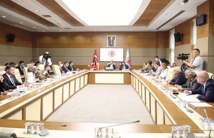 Görselde, Türkiye Büyük Millet Meclisinde gerçekleştirilen komisyon toplantısı sırasında milletvekilleri dikdörtgen biçimindeki bir masanın etrafında oturuyor. Komisyon Başkanı Mehmet Muharrem Kasapoğlu, kameranın çekim açısına göre masanın tam karşı tarafında yer alıyor.