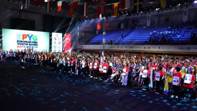 Görselde büyük bir spor salonunda düzenlenen uluslararası bir açılış töreni görülüyor. Sahnenin solunda dev ekranda “EPYG European Para Youth Games” logosu yer alıyor. Alanda farklı ülkelerden gelen sporcular ellerinde ülke bayrakları ve pankartlarla sıraya dizilmiş. Üst kısımda çeşitli ülke bayrakları salonu süslüyor. Katılımcılar arasında tekerlekli sandalye kullanan sporcular da var. Bu sahne, 2025 Avrupa Para Gençlik Oyunları’nın coşkulu açılış anını yansıtıyor.