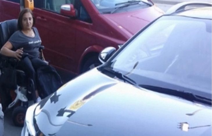 Görselde akülü tekerlekli sandalye kullanan bir kadın kaldırımda duruyor. Önünde ve yanında park edilmiş araçlar (siyah ve kırmızı renkli) bulunuyor. Araçların kapattığı alan nedeniyle kadının geçişi engellenmiş görünüyor. Bu durum erişim sorunu yaşandığını gösteriyor.