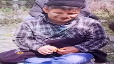 Fotoğrafta, açık havada çekilmiş bir karede, kareli gömlek, mavi kot pantolon ve siyah bere takan bir Tunay Çetiner oturuyor. Kucağında koyu renk bir çanta bulunuyor ve ellerinde küçük bir nesne tutuyor. Arka planda bulanık şekilde başka insanlar ve doğa manzarası görünüyor.