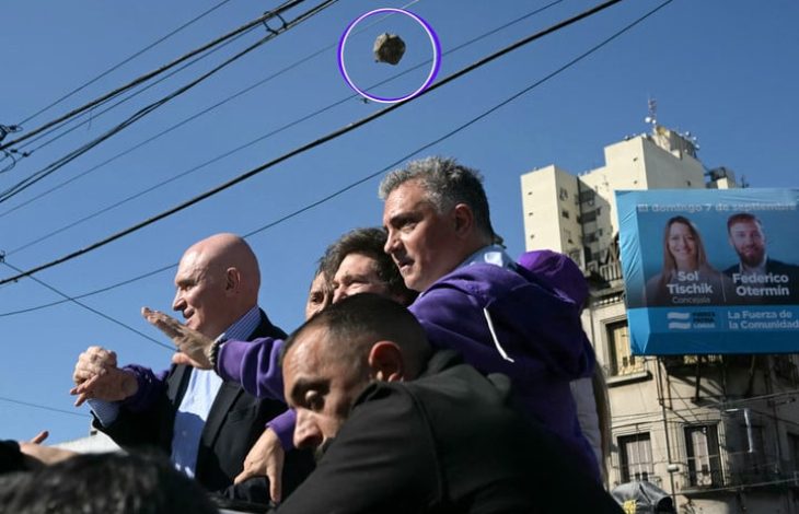 Görselde, kalabalık bir ortamda siyasetçi ya da yetkili konumunda olduğu anlaşılan bir grup insan yer alıyor. Ön planda birkaç erkek şahıs dikkatlice etrafa bakarken yukarıdan bir taşın havada uçmakta olduğu an objektife yansımış. Taş mor bir daire ile işaretlenmiş. Arka planda apartmanlar, elektrik telleri ve seçim afişleri görünüyor. Afişte “Sol Tischik” ve “Federico Otermin” isimleri okunabiliyor. Görsel, Arjantin Devlet Başkanı Javier Milei’nin seçim kampanyası sırasında taşlı saldırıya uğradığı anı belgeleyen bir fotoğraf niteliğinde.