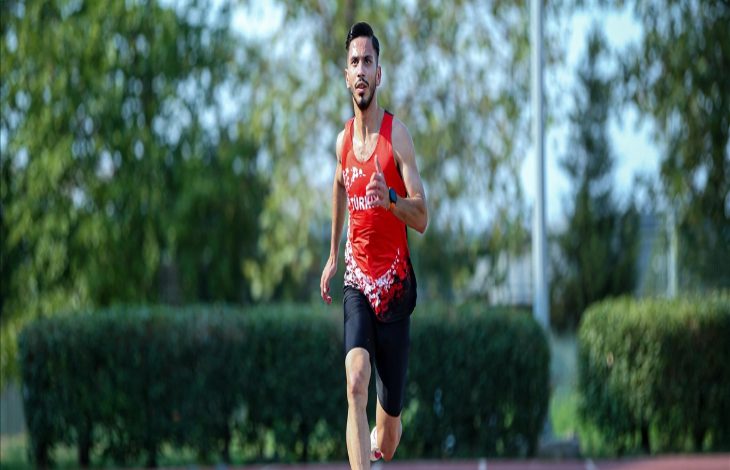 Görselde, kırmızı formasıyla pistte koşan atlet Çağatay Çelik görülüyor. Arka planda yeşil çalılar ve ağaçlar yer alıyor, sporcu odaklı ve hareket hâlinde çekilmiş bir fotoğraf.