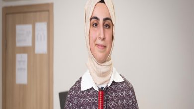 Görselde, başörtülü genç bir kadın Ayşe Begüm Başbozkurt Gür iç mekânda kameraya doğru gülümseyerek poz veriyor. Üzerinde desenli bir tunik ve beyaz yakalı bir gömlek bulunuyor. Boynunda bir mikrofon ya da ses cihazı klipsi var, bu da bir röportaj ya da tanıtım çekimi olabileceğini düşündürüyor. Arka planda kapalı bir kapı ve duvarda bazı kağıtlar asılı.