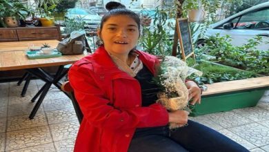 Görselde, genç bir kadın bir kafede oturuyor. Üzerinde kırmızı bir mont, koyu renk pantolon ve siyah üst var. Elinde beyaz çiçeklerden oluşan bir buket tutuyor. Arka planda yeşil bitkiler, ahşap masa-sandalyeler ve camın dışından görünen park halindeki bir araç yer alıyor.