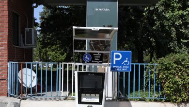 Bir bina önünde, Diyarbakır Büyükşehir Belediyesi'ne ait "Engelli Araç Şarj İstasyonu" ve "Engelli Araç Park Yeri" tabelalarının bulunduğu bir kiosk görülüyor. Kioskun üstünde güneşlik, arkasında ise ağaçlar ve bir çit yer alıyor.