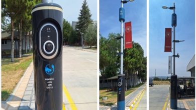 Görselde, bir sokak üzerinde konumlandırılmış modern teknolojiyle donatılmış aydınlatma direkleri görülüyor. Direklerin alt kısmında “Elektrikli Araç Şarj Ünitesi” yer almakta ve Togg logosu dikkat çekiyor. Direklerin üst bölümlerinde ise sokak lambaları ve çeşitli sensörler bulunuyor. Ayrıca direklere Enerji ve Tabii Kaynaklar Bakanlığı'nı temsil eden kırmızı bayraklar asılmış.