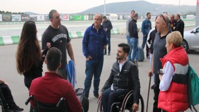 Görselde, çoğunluğu tekerlekli sandalye kullanan ve baston destekli bireylerden oluşan bir grup, açık havada pist kenarında sohbet ediyor. Arka planda pist, bariyerler ve uzaklarda dağ manzarası görülüyor.