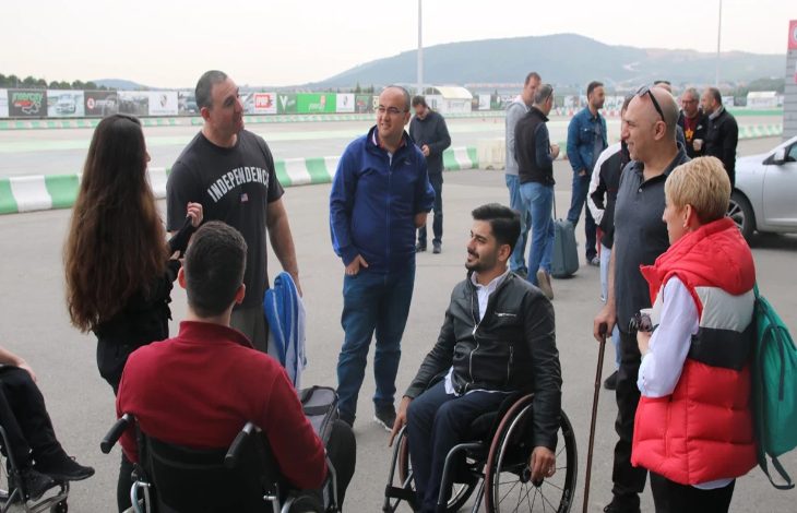 Görselde, çoğunluğu tekerlekli sandalye kullanan ve baston destekli bireylerden oluşan bir grup, açık havada pist kenarında sohbet ediyor. Arka planda pist, bariyerler ve uzaklarda dağ manzarası görülüyor.