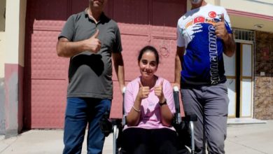 Görselde, açık havada çekilmiş bir karede üç kişi yer alıyor. Ortadaki genç kadın akülü tekerlekli sandalyede oturuyor ve iki baş parmağını yukarı kaldırarak gülümsüyor. Yanında ayakta duran iki erkek de aynı şekilde baş parmaklarını yukarı kaldırmış. Hepsi mutlu ve gururlu görünüyor. Arka planda bir bina girişi ve kapalı bir garaj kapısı dikkat çekiyor.