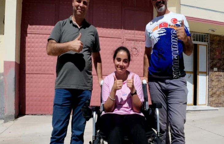 Görselde, açık havada çekilmiş bir karede üç kişi yer alıyor. Ortadaki genç kadın akülü tekerlekli sandalyede oturuyor ve iki baş parmağını yukarı kaldırarak gülümsüyor. Yanında ayakta duran iki erkek de aynı şekilde baş parmaklarını yukarı kaldırmış. Hepsi mutlu ve gururlu görünüyor. Arka planda bir bina girişi ve kapalı bir garaj kapısı dikkat çekiyor.