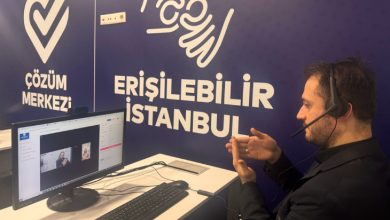 Fotoğrafta, bir erkek bilgisayar başında oturuyor ve kulaklık-mikrofon seti takılı. Ekranda çevrim içi bir görüşme gerçekleştiriliyor. Erkek kişi, ellerini kullanarak işaret diliyle iletişim kuruyor. Duvarın arkasında beyaz renkte büyük harflerle "ERİŞİLEBİLİR İSTANBUL" ve "ÇÖZÜM MERKEZİ" yazıları yer alıyor. Aynı zamanda işaret diliyle "erişilebilir" kelimesini temsil eden bir simge de duvarda bulunuyor. Masa üzerinde bir bilgisayar monitörü, klavye, fare ve cep telefonu yer alıyor. Görsel, İstanbul'da engelliler için erişilebilirlik ve iletişim destek hizmetleri sağlandığını gösteren bir ortamı yansıtıyor.