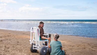 Görselde, sahilde bir erkek ve bir kadın denize karşı oturuyor. Erkek, geniş tekerlekli, kumda hareket edebilen bir plaj tekerlekli sandalyesinde; kadın ise kumun üzerine oturmuş. İkisi el ele tutuşarak huzurlu bir şekilde denizi izliyor. Arka planda denize giren insanlar ve ufukta şehir silueti görünüyor.