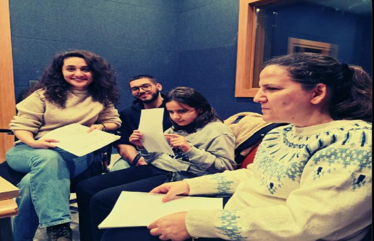 Görselde dört kişi bir stüdyo ortamında oturuyor. Üçü ellerinde kağıt tutuyor; biri gülümserken, diğerleri ciddi bir şekilde belgeleri inceliyor. Arkada ses yalıtımı yapılmış bir duvar ve stüdyo camı görülüyor. Radyo ya da ses kaydı ortamında oldukları izlenimi veriyor.