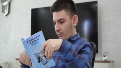 Görselde genç bir erkek Mert Esat Ölmez, mavi kareli bir gömlek giymiş şekilde elindeki açık kitabı dikkatle okuyor. Kitabın kapağında “Zorluklardan Güç Almak” yazıyor. Arka planda televizyon ve ev ortamına ait eşyalar görünüyor.