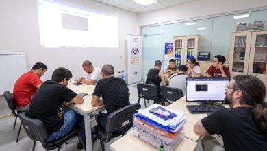 Katılımcılar, iki ayrı masa etrafında oturmuş durumda ve çoğunlukla tablet veya defter gibi materyallerle meşguller. Arka plandaki duvarda bir projeksiyon ekranına bir video yansıtılıyor. Sağda, "Destekli İstihdam Programı" yazılı bir tanıtım afişi bulunmakta. Arka planda cam bölmelerle ayrılmış bir alan ve kitaplıklar yer almakta. Ön planda ise bir eğitmen veya görevli, bilgisayar ekranından projeksiyondaki video ile ilgili bir şeyi kontrol ediyor.