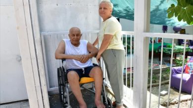 Görselde, açık hava ortamında tekerlekli sandalyede oturan yaşlı bir adam ve yanında ayakta duran bir kadın yer alıyor. Adam beyaz atlet ve koyu şort giymiş, çıplak ayaklı. Kadın ise açık renkli bir tişört ve pantolon giymekte, turuncu terlikleri var. İkili bir evin girişinde konumlanmış; etraflarında beyaz parmaklıklar bulunuyor. Arka planda yeşil bir tente ve renkli minderlerle dekore edilmiş bir bahçe alanı dikkat çekiyor.