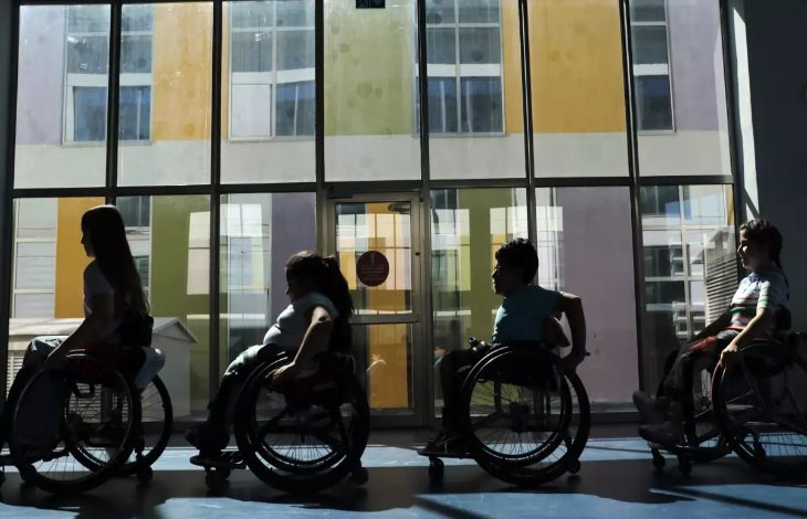 Görselde, tekerlekli sandalyedeki dört çocuk yan yana dizilmiş şekilde kapalı bir alanda görülüyor. Arkadan gelen ışık nedeniyle silüetleri öne çıkıyor. Büyük camlı kapının önünde, renkli duvarlarla çevrili bir binanın manzarası dikkat çekiyor.