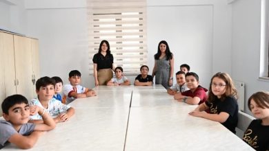 Görselde sınıf ortamında, dikdörtgen masaların etrafına oturmuş çocuklar yer alıyor. Masanın iki ucunda ise ayakta duran iki kadın bulunuyor. Çocuklar kameraya doğru bakıyor, sınıf düzeninde bir etkinlik havası var. Arka planda pencere ve kapalı stor perde görünüyor.