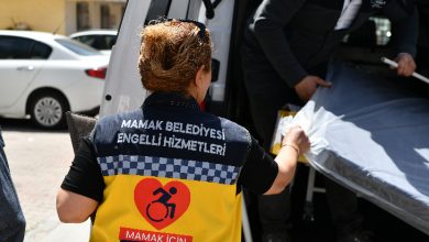Görselde, sırtında “Mamak Belediyesi Engelli Hizmetleri” yazan bir yelek giymiş görevli, bir araca eşya (muhtemelen yatak veya medikal malzeme) yerleştirmeye yardım ediyor. Yeleğin üzerinde tekerlekli sandalye sembolü ve “Mamak için canla başla” ifadesi bulunuyor. Arka planda başka bir görevli ve park etmiş araçlar görünüyor.