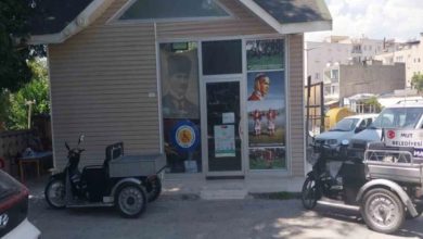 Fotoğrafta küçük, tek katlı bir bina görülüyor. Binanın dış cephesi ahşap kaplama ve üçgen çatıya sahip. Giriş kapısı camdan yapılmış ve kapının her iki yanında cam paneller bulunuyor. Sol taraftaki camın arkasında büyük bir Atatürk posteri asılı. Girişin hemen önünde tekerlekli, motorlu engelli aracı park etmiş. Sağ tarafta ise “Mut Belediyesi” yazılı başka bir motorlu araç bulunuyor.