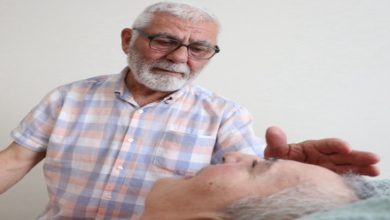 Fotoğrafta, beyaz saçlı ve sakallı, gözlüklü yaşlı bir erkek, yatakta yatan beyaz saçlı bir kadına sevgi dolu bir ifadeyle bakıyor ve elini şefkatle başına doğru uzatıyor. Arka plan sade bir duvardan oluşuyor.