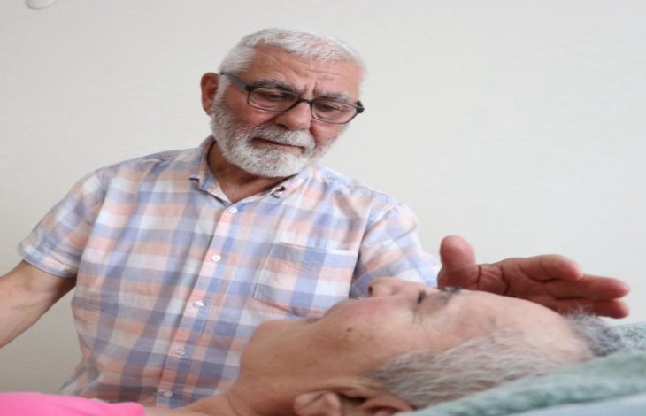 Fotoğrafta, beyaz saçlı ve sakallı, gözlüklü yaşlı bir erkek, yatakta yatan beyaz saçlı bir kadına sevgi dolu bir ifadeyle bakıyor ve elini şefkatle başına doğru uzatıyor. Arka plan sade bir duvardan oluşuyor.