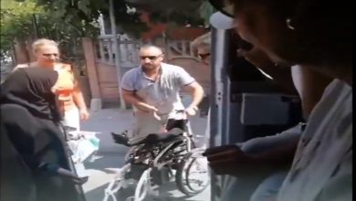 Görselde, dışarıda duran bir adam katlanmış bir tekerlekli sandalyeyi tutarken, çevresinde birkaç kişi bulunuyor. Fotoğraf, bir aracın içinden dışarıya doğru çekilmiş ve sokak ortamı net şekilde görülüyor.