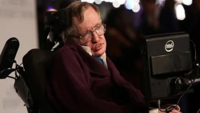 Görselde, ünlü teorik fizikçi Stephen Hawking yer alıyor. Elektrikli tekerlekli sandalyesinde oturmakta ve yanında, iletişim kurmak için kullandığı Intel destekli özel bilgisayar ekranı bulunuyor. Üzerinde koyu renk bir giysi var ve yüz ifadesi ciddi. Arka planda flu şekilde ışıklar ve insanlar görünüyor, bu da etkinlik tarzı bir ortam izlenimi veriyor.