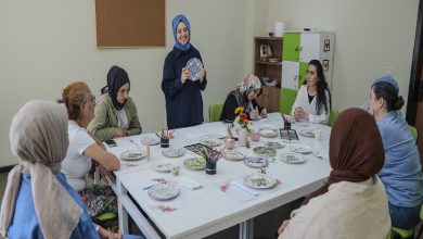 Görselde bir grup kadın masa etrafında oturmuş atölye çalışması yapıyor. Masanın üzerinde boyama kalemleri, fırçalar ve desenli seramik tabaklar yer alıyor. Ayakta duran bir kadın, elindeki tabakla diğerlerine bir şeyler anlatıyor ve gülümseyerek sunum yapıyor.