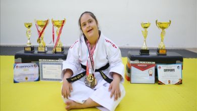 Görselde, karate kıyafeti giymiş genç bir kız yerde oturuyor. Boynunda kazandığı madalyalar asılı. Arka planda ise çok sayıda kupa ve başarı sertifikası sıralanmış. Kızın gülümseyen ifadesi, spor yoluyla elde ettiği başarı ve gururu yansıtıyor.