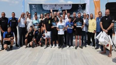 Görselde açık havada bir grup insan toplu fotoğraf çekiliyor. Ön sırada ellerinde sertifika tutan engelli sporcular ve yanlarında antrenörler bulunuyor. Arka planda etkinlik afişi ve bayraklar yer alıyor. Katılımcıların çoğu spor kıyafetli, bazılarının dalış kıyafeti giydiği görülüyor.