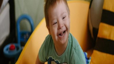 Görselde, yeşil tişört giymiş küçük bir çocuk gülümseyerek mutlu bir şekilde poz veriyor. Arkada oyuncakların bulunduğu bir ortam ve sarı bir koltuk ya da kaydırak benzeri bir eşya dikkat çekiyor. Çocuğun neşeli ifadesi, samimi ve sıcak bir atmosfer yansıtıyor.