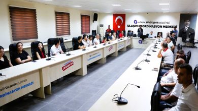 Antalya Büyükşehir Belediyesi Ulaşım Koordinasyon Merkezi’ne ait geniş bir toplantı salonunda gerçekleşen bir toplantı görülüyor. U şeklinde dizilmiş uzun masaların etrafında farklı yaş ve cinsiyetlerden katılımcılar yer alıyor. Sol ve sağ tarafta katılımcılar otururken, salonun ön tarafında bir konuşmacı ya da yönetici bulunuyor.