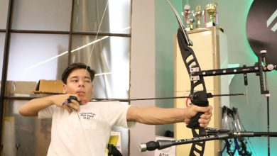 Görselde, genç bir erkek sporcu okçuluk yaparken görülüyor. Beyaz tişört giymiş ve yayı germiş halde hedefe odaklanıyor. Arkada kupa dolabı ve “Atış Çizgisi” yazılı tabela dikkat çekiyor. Spor salonu ortamında çekilmiş bir fotoğraf.