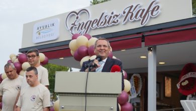 Ayvalık Belediye Başkanı Mesut Ergin, Engelsiz Kafe önünde kürsüde konuşma yaparken yanında down sendromlu üç erkek personel bulunuyor.