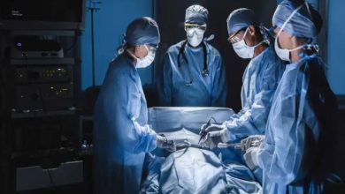 Görselde ameliyathane ortamında maske ve mavi ameliyat kıyafetleri giymiş cerrahlar bir operasyon gerçekleştiriyor. Masada üzeri örtülü bir hasta yatmakta, doktorlar odaklı şekilde çalışıyor. Arka planda bir başka doktor izliyor.