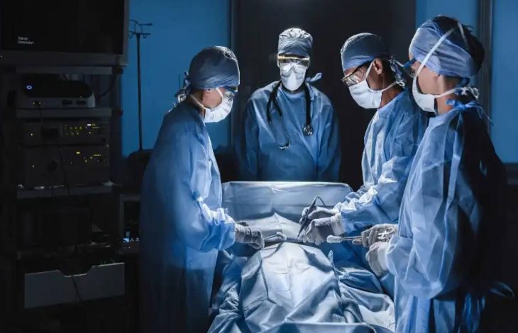 Görselde ameliyathane ortamında maske ve mavi ameliyat kıyafetleri giymiş cerrahlar bir operasyon gerçekleştiriyor. Masada üzeri örtülü bir hasta yatmakta, doktorlar odaklı şekilde çalışıyor. Arka planda bir başka doktor izliyor.