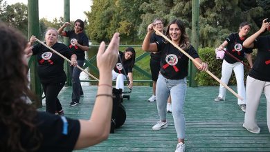Görselde, bir grup kadın açık havada ahşap zeminde kürek antrenmanı yapıyor. Kadınlar siyah tişört giymiş ve ellerinde uzun küreklerle çalışıyor. Önde yer alan kadın gülümseyerek kameraya bakıyor. Arkada ağaçlar ve yeşil bir doğa manzarası bulunuyor. Fotoğraf, takım ruhu ve enerji dolu bir anı yansıtıyor.