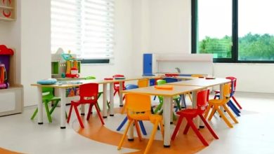 Görselde renkli ve modern bir çocuk sınıfı görülüyor. Masaların etrafında kırmızı, sarı, yeşil ve mavi sandalyeler yer alıyor. Ortam geniş, ferah ve aydınlık; büyük pencerelerden dışarıdaki yeşillik görünüyor. Çocuklara yönelik eğitim ve oyun alanı izlenimi veriyor.